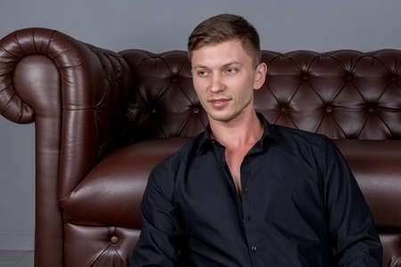 Sexy handsome man wearing black shirt sitting on floor near the leather sofaの写真素材