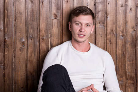 Young handsome man wearing white sweater sitting near wood wall