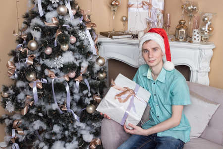 Young man with big present sitting on couch near christmas tree and fireplaceの写真素材