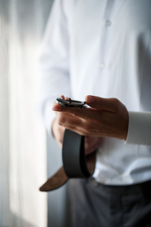Elegant businessman man fastening a belt on his trousers he is wearing a white dress shirt