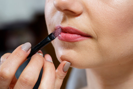 Makeup artist applies red lipstick. Beautiful female face. Hand of a make-up master painting lips of a young girl beauty model. Makeup in progressの写真素材