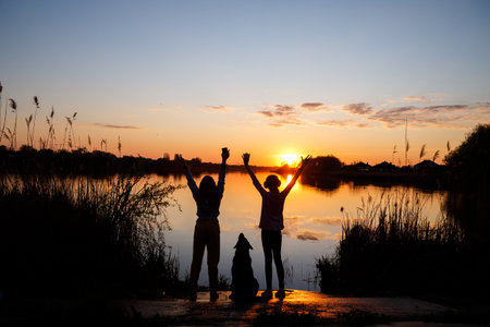 Silhouettes of girls with a dog at sunset on the lake, the sun sets behind the trees and beautiful reflections in the waterの写真素材