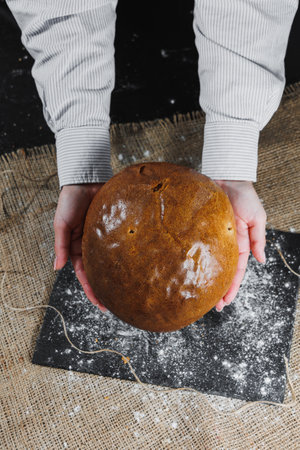 Homemade crispy loaf of bread. Baker holds fresh bread in his hands. Dark background