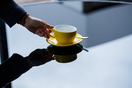 Yellow cup for coffee or tea with a saucer in the hands of a girl on the background of a glass...