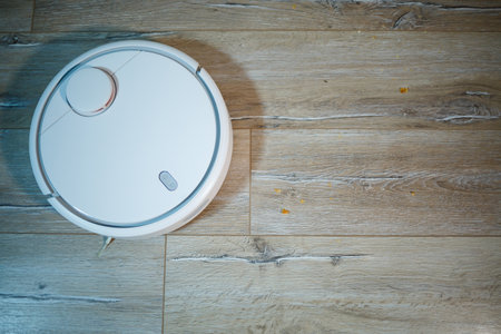 A girl sitting at home on the sofa eating a croissant and drinking tea. Robot vacuum cleaner removes crumbs from food on the floor. Simple and easy cleaning with the use of modern technology.の写真素材