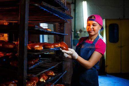 Girl baker holds fresh pies in the hands of the bakery. She is wearing a denim frock and a cap. Production of bakery products. Rack with hot crispy pastries.の写真素材
