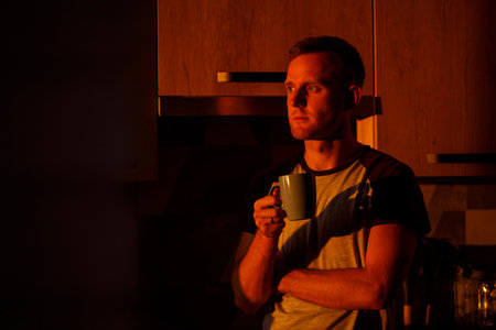 A man standing in front of a window in sunset lighting is drinking coffee in his kitchen