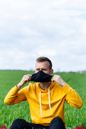 Young man sitting on green grass in a protective antiviral mask and thinking about the future. The guy is resting in the park on a sunny summer day. The concept of rest. Virus protection. Quarantineの写真素材