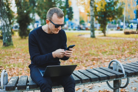 Attractive young man in a black sweater and sunglasses, he is sitting in an autumn park on a bench and works on a laptopの写真素材