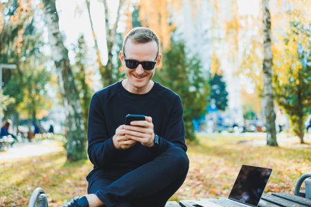 Cute successful young man in sunglasses sitting on park bench with laptop and phone and looking away on sunny day. Freelance, remote work concept.の写真素材