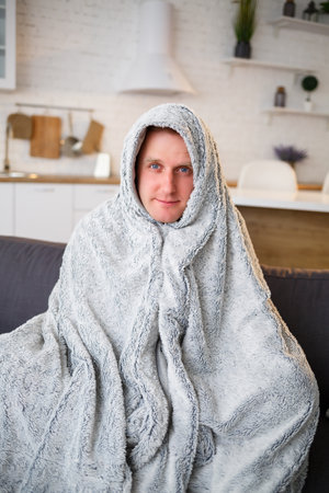 A sick man sits at home on a gray sofa with a white blanket. Illness, protection, coronavirus, illness, flu.の写真素材