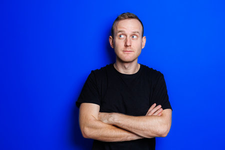 A young man in a black T-shirt on a blue background shows different emotions on his face. Cheerful man with a smile. Place for text. Male beautyの写真素材