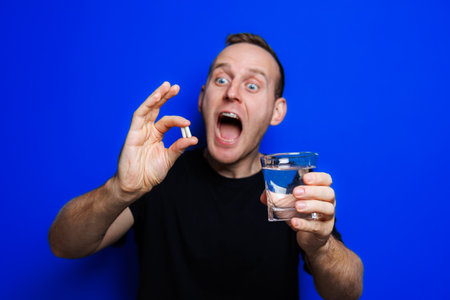 A young man in a black T-shirt on a blue background stands with a thermometer in his hands. An increase in body temperature, feeling sick. Weakened immunity. Selective focusの写真素材