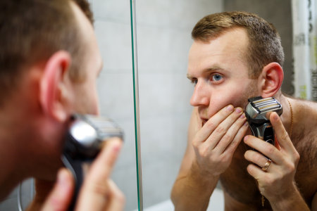 A man shaves his face with an electric razor in front of a mirror. Skin irritation. Bath procedureの写真素材