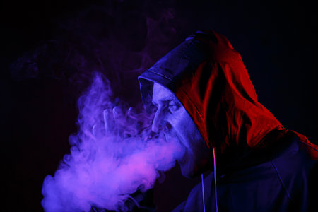 A man in a protective suit exhales smoke. Halloween image concept. Virus protection. Illuminated with colored lightsの写真素材
