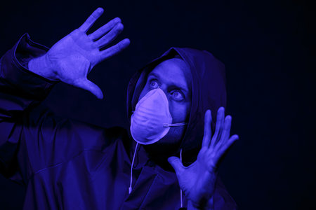 A man in a protective suit and white mask in a dark room. Halloween image concept. Virus protection. Illuminated with colored lightsの写真素材