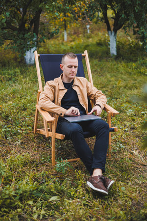 A handsome man in a brown shirt sits on a wooden folding chair in the garden and works with a laptop. Wooden ecological furniture. Remote workの写真素材
