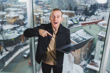 Portrait of a young joyful man with a laptop in his hands, standing near the window. Work in the...