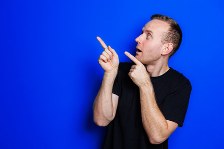 A young man in a black T-shirt on a blue background shows different emotions on his face. Cheerful man with a smile. Place for text. Male beautyの写真素材