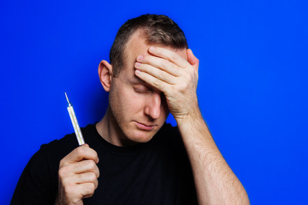 A young man in a black T-shirt on a blue background stands with a thermometer in his hands. An...