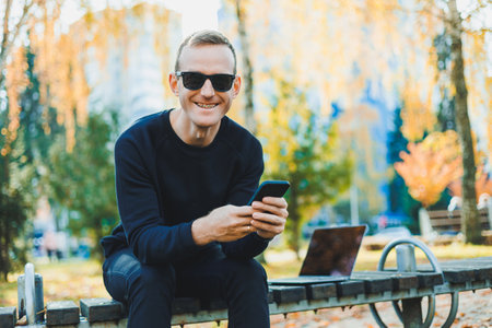 Cute successful young man in sunglasses sitting on park bench with laptop and phone and looking away on sunny day. Freelance, remote work concept.の写真素材