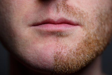 Young man shaving his beard. He shaved off half the bristles against a gray background. Male face care. Shaving bristles. The difference between skin without hair and with a mustacheの写真素材