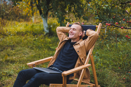 A young man works on a laptop outdoors. A young freelancer rests in the forest. Remote work, active recreation in the summer. Tourism, people concept - man sitting on a chair outdoors.の写真素材