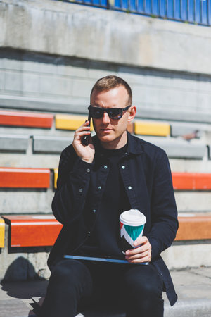 Photo of smiling businessman in suit holding mobile phone while standing outdoor near building with...