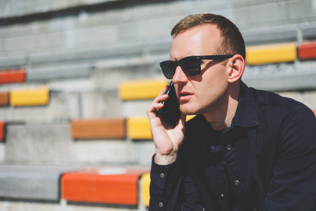 Cheerful hipster guy in trendy outfit smiling while resting at urban setting in city using cellphone device on leisure, happy Middle Eastern male blogger in sunglasses enjoying travel lifestyleの写真素材