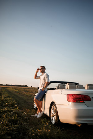 Young successful male businessman is standing near his white convertible car. A guy in sunglasses in a white shirt and shorts near his car.の写真素材