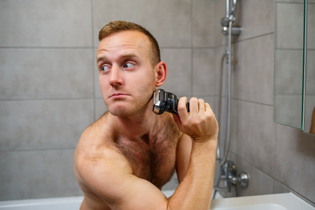 A man shaves his face with an electric razor in front of a mirror. Skin irritation. Bath procedureの写真素材
