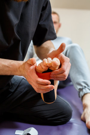Male physiotherapist doctor massages the feet of a relaxed man sitting on a stretcher. He uses kinesio tape. Taping the footの写真素材