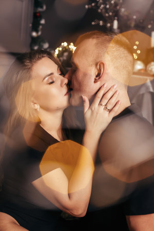 View of a passionate romantic couple spending time before New Year at home with garlands. Young woman and handsome man hug, kiss. Christmas time.の写真素材