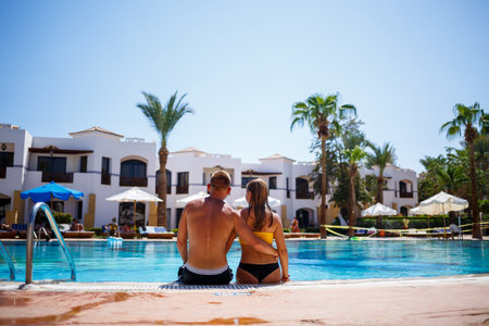 Happy man guy joy on vacation, girl sitting next to in a yellow swimsuit. Large pool, palm trees, blue water in the poolの写真素材