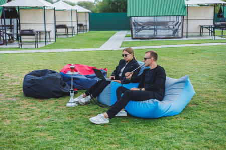 A young couple relaxes in the park and smokes hookah. A man and a woman sit on chairs in nature and smoke a hookah. Family vacationの写真素材