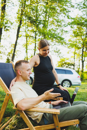 A happy man and his pregnant wife are working on a laptop while sitting in nature. A young couple is relaxing in nature and watching a movie on a laptop.の写真素材