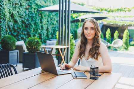 Young positive female freelancer sitting at a table with a laptop and working on a green terrace, looking at the screen and working on a project remotely in a cafeの写真素材