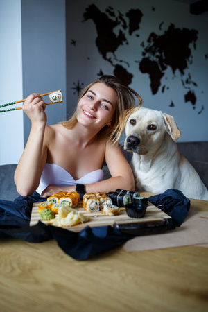 Beautiful girl eating sushi close-up with her dog labrador, smiling, holding a sushi roll with chopsticks. Healthy Japanese food.の写真素材