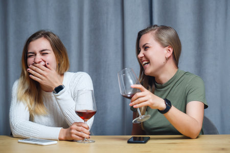 two girls of European appearance with blond hair are sitting at the table, drinking wine and laughing, relaxing at home, alcoholの写真素材