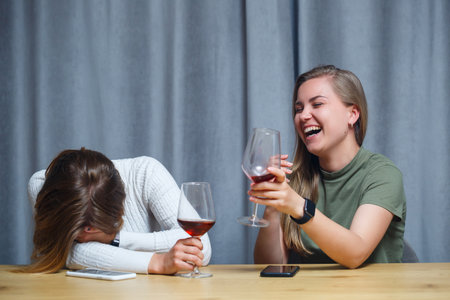 two girls of European appearance with blond hair are sitting at the table, drinking wine and laughing, relaxing at home, alcoholの写真素材