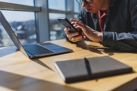 Cropped image of man holding smartphone, receiving transaction confirmation message on laptop...