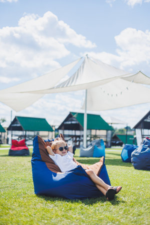 A fair-haired boy of 5-6 years old in a white T-shirt and shorts is sitting on a bean bag chair. A child in sunglasses rests in the fresh airの写真素材