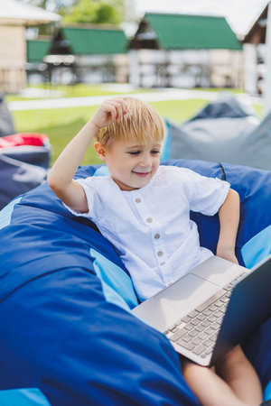 Little boy with a laptop on the background of summer grass. A boy in a white T-shirt is sitting on a beanbag in the park.の写真素材