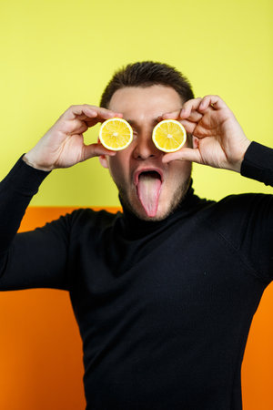 Portrait of a man on a yellow background in black raglan with cut lemon instead of eyes. A guy with lemons near his face. A smile on his face. An image for a male photo. Photo session conceptの写真素材