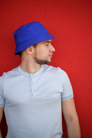 Young handsome man with a beard in a panama hat on his head against the background of a red wall....