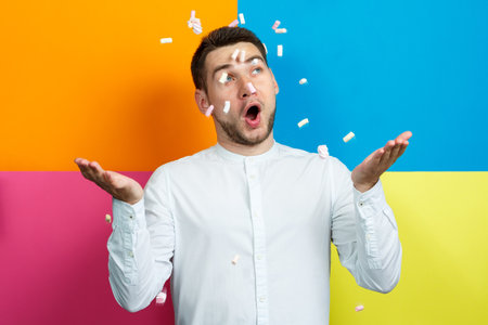Marshmallows sprinkle on the mans head. Bright man portrait with sweets on his hair. Sweets on the guy's face. Cheerful man portrait with sweets. Funny emotionsの写真素材
