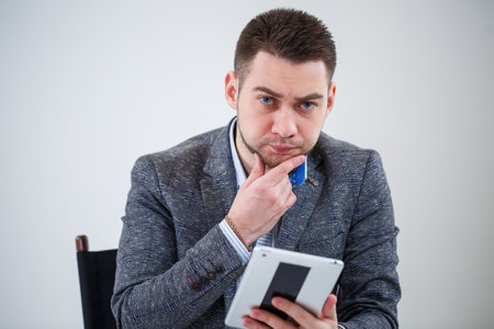 Successful business man businessman in a suit sitting on a wooden chair with a tablet and looking at the screen. On a white background with thoughts of a new projectの写真素材