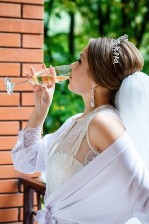 Beautiful bride in a white coat on the balcony drinks champagneの写真素材