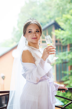 Beautiful bride in a white coat on the balcony drinks champagneの写真素材