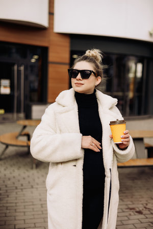 A young pretty woman in a white fur coat and sunglasses with coffee walks around the city. Stylish young girl.の写真素材